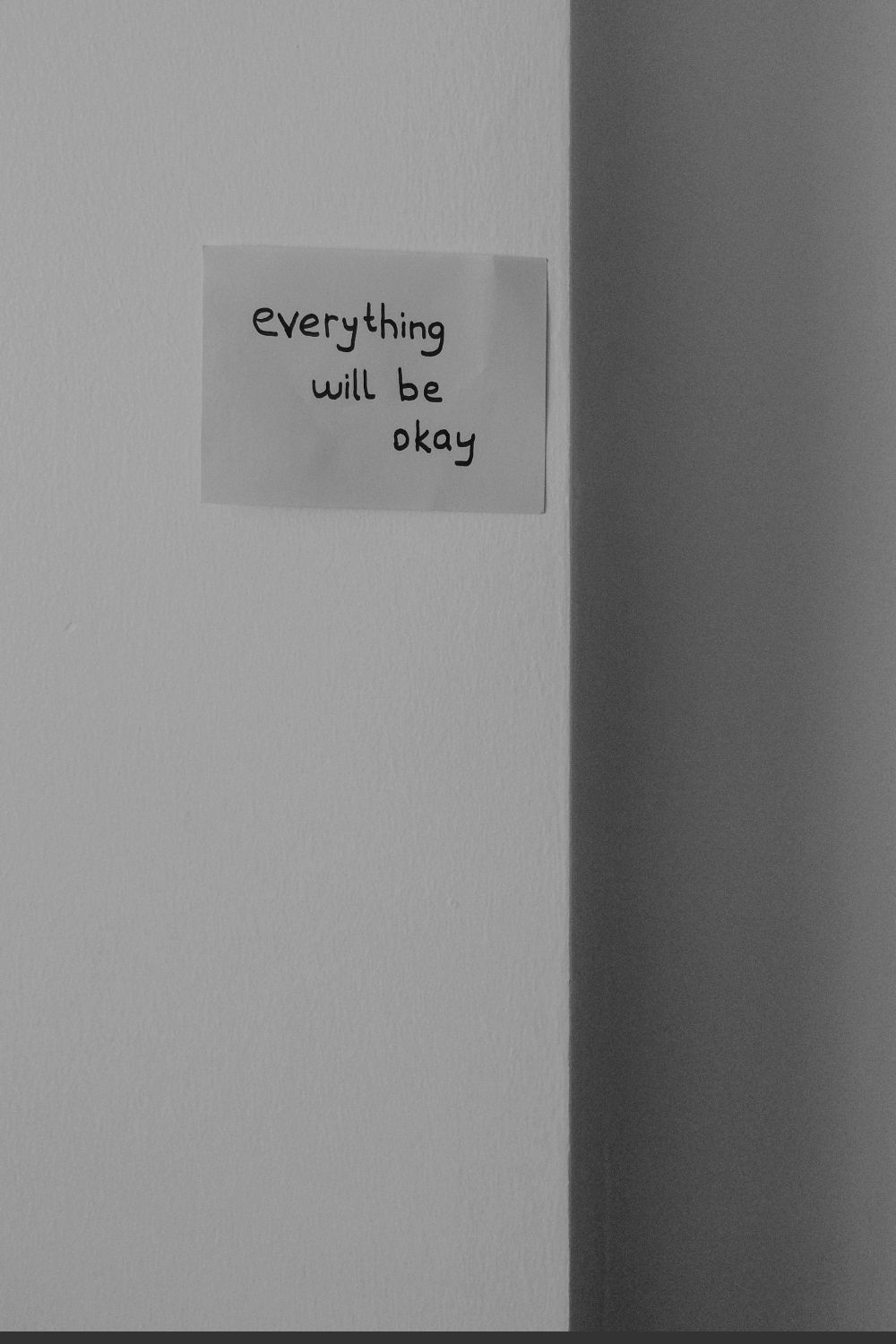 A handwritten note on white paper affixed to a wall reads "everything will be okay" in black ink, positioned against a minimalist background with stark shadows.
